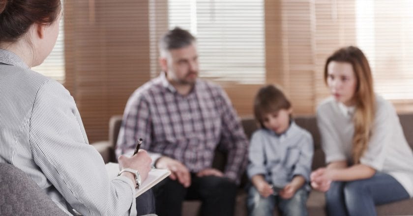 Rear view of female psychologist helping young family with a kid to solve child development problems. Family sitting on a sofa in the blurred background